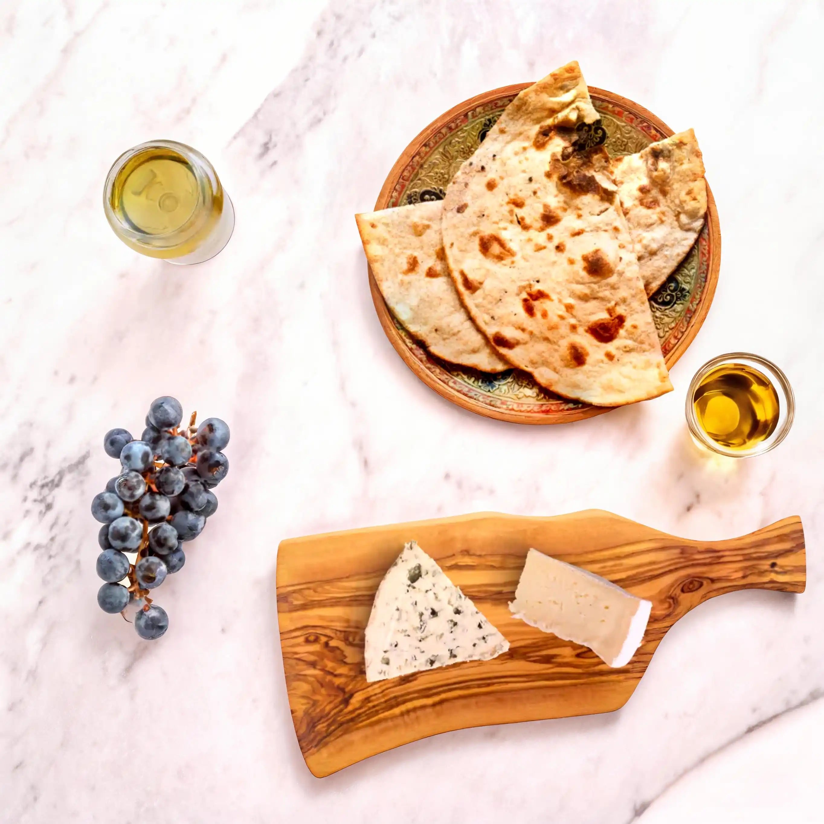 Olive wood serving board styled with grapes, fresh bread, cheese, and a glass of wine, highlighting its natural grain and rustic charm in an elegant dining arrangement.